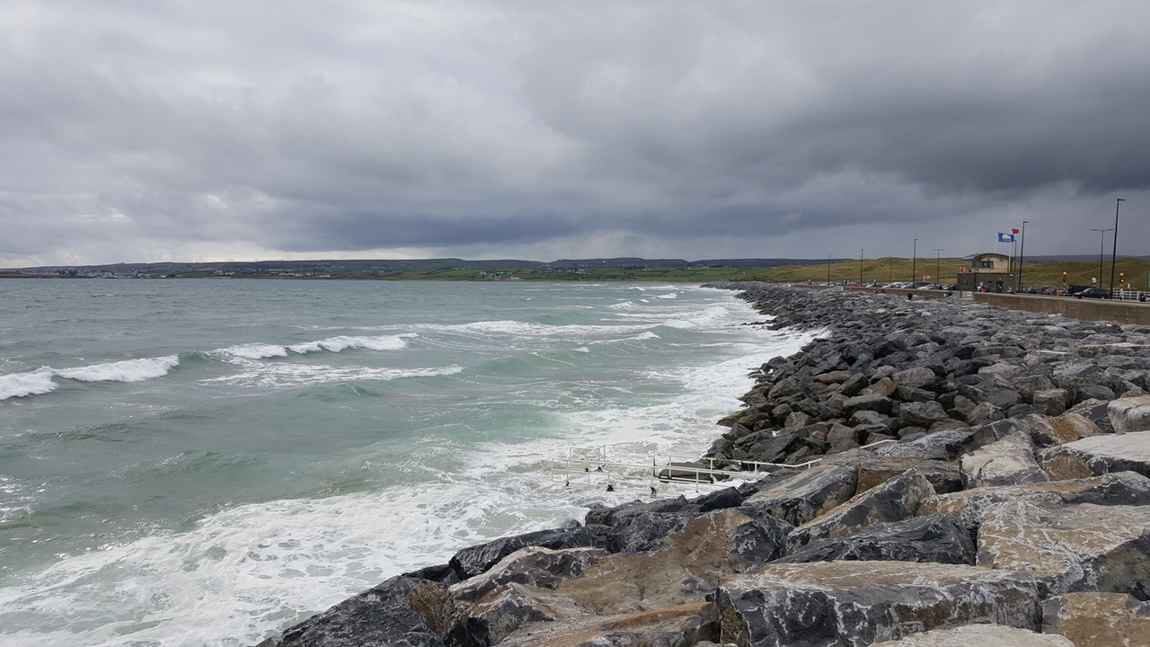 Une côte rocheuse avec des vagues qui se brisent, sous un ciel nuageux et qui s'assombrit.