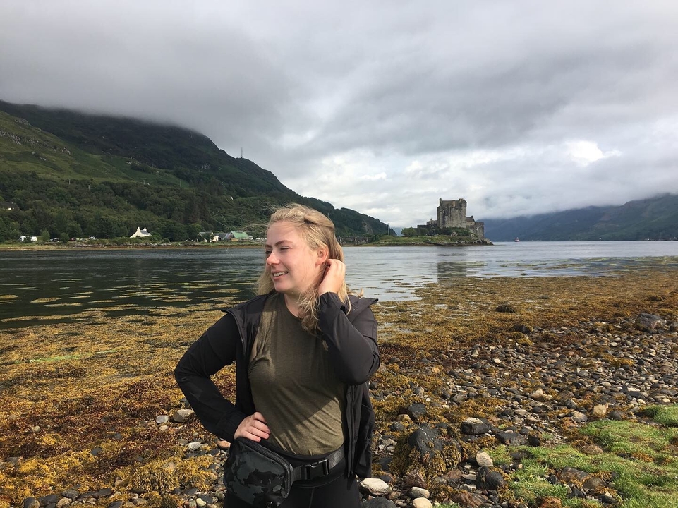 Person standing near a scenic waterfront with a castle in the background.