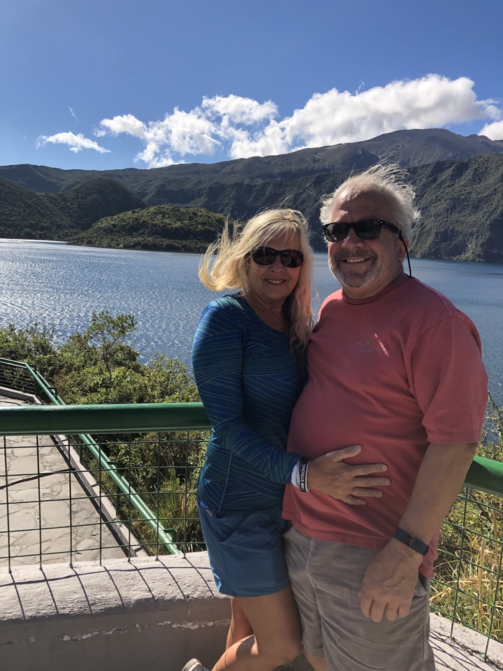 Couple souriant au bord d'un lac avec des montagnes en arrière-plan.