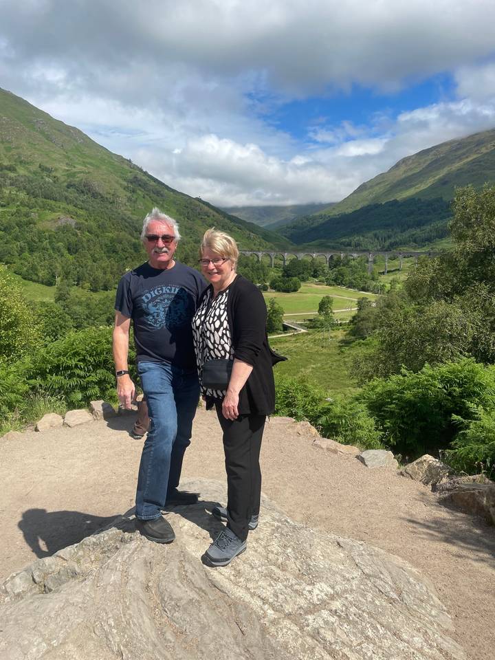 Two people standing on a rocky surface with a scenic view of hills and sky.