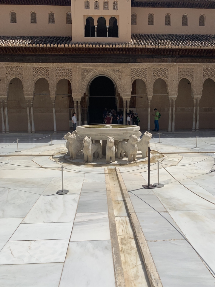 Ornate stone fountain surrounded by arches with visitors.