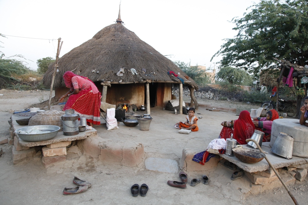 Scène rurale avec une hutte traditionnelle, des femmes qui cuisinent dehors et un enfant assis.