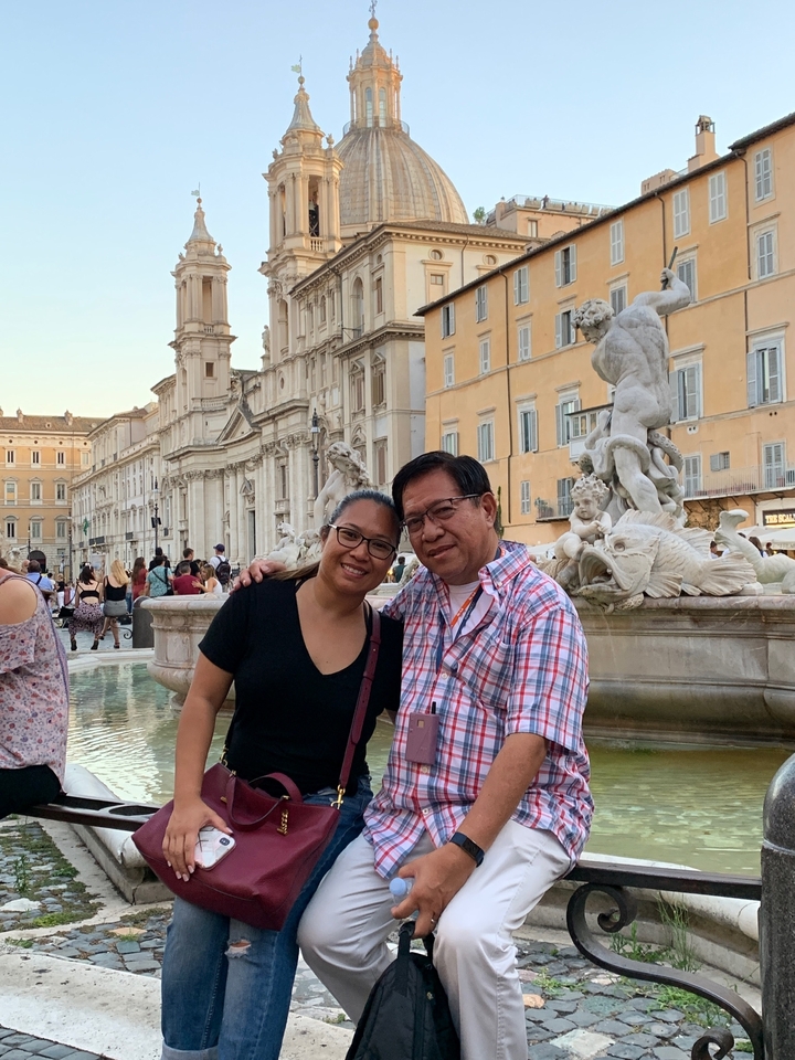 Couple posant devant une fontaine avec des bâtiments historiques en arrière-plan.