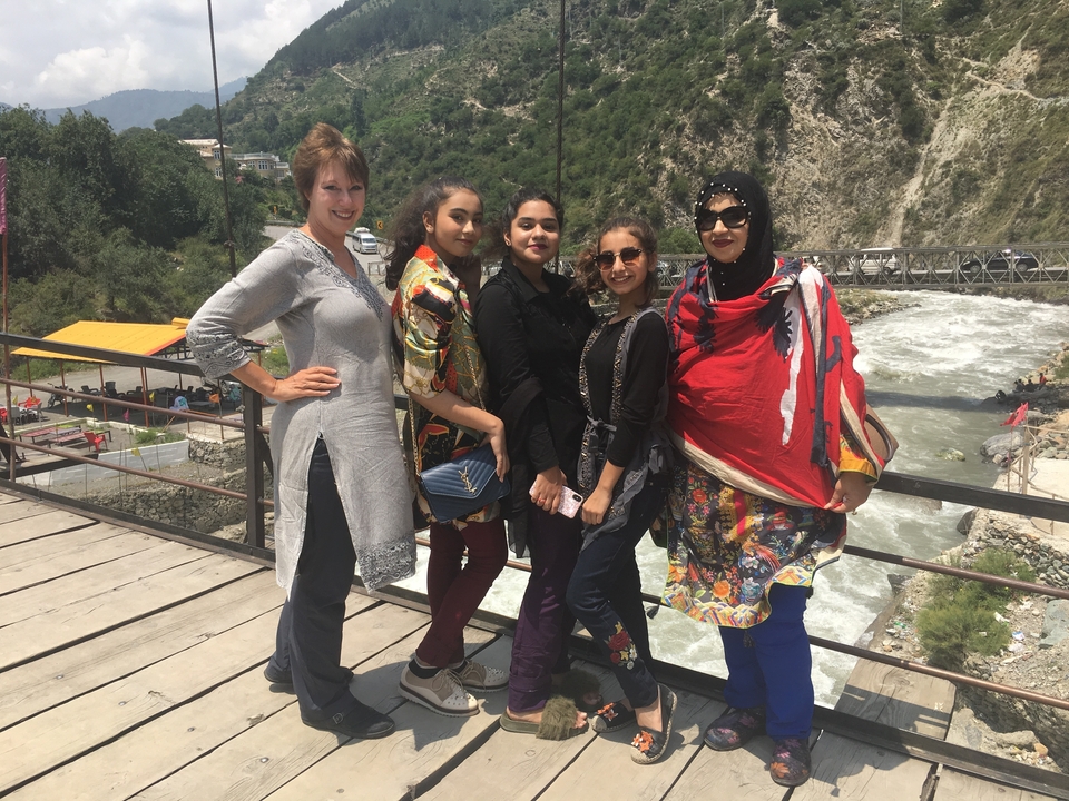 Cinq femmes posant sur un pont avec une rivière et des collines en arrière-plan.