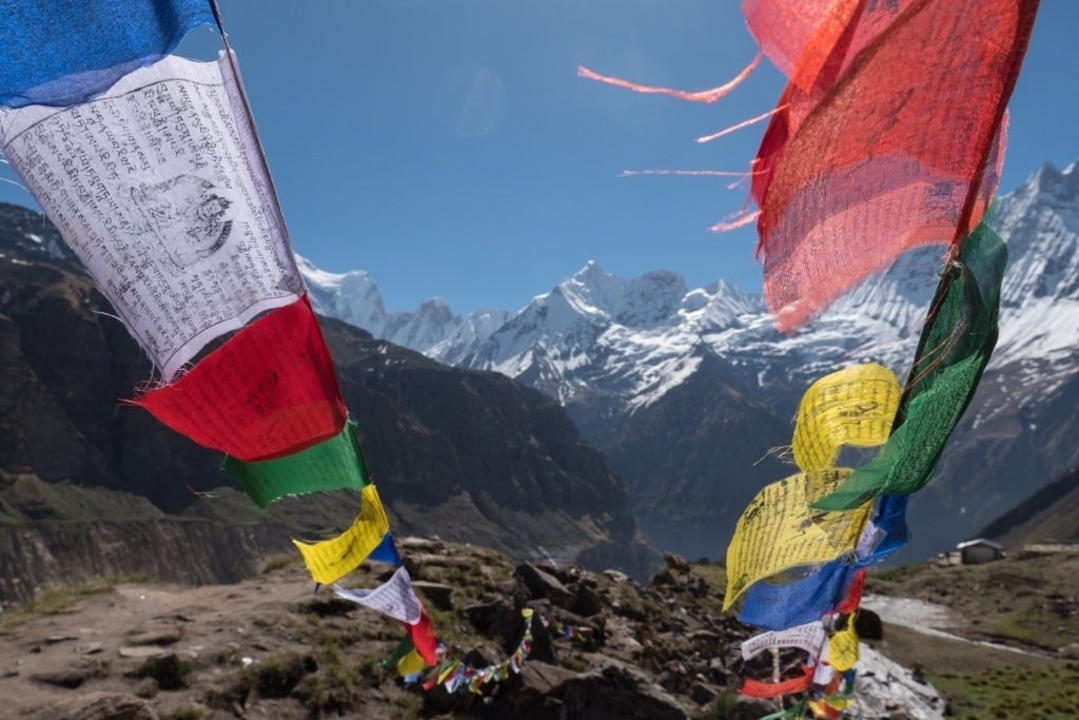Colorful Buddhist prayer flags fluttering in foreground with snow-capped Annapurna peaks and deep blue sky beyond.