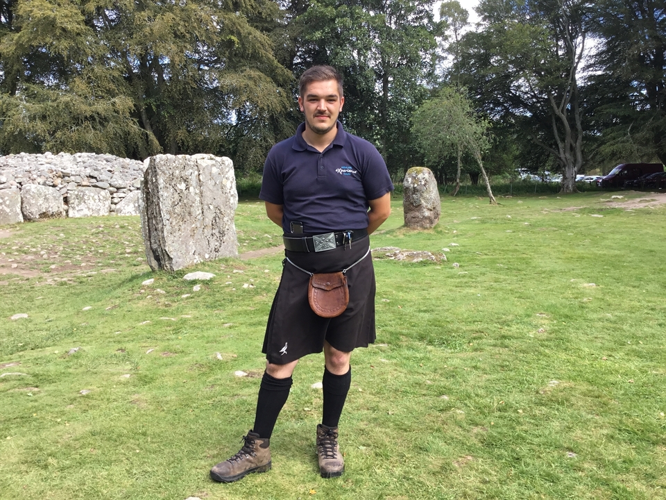 Homme en vêtements traditionnels debout dans une zone herbeuse.