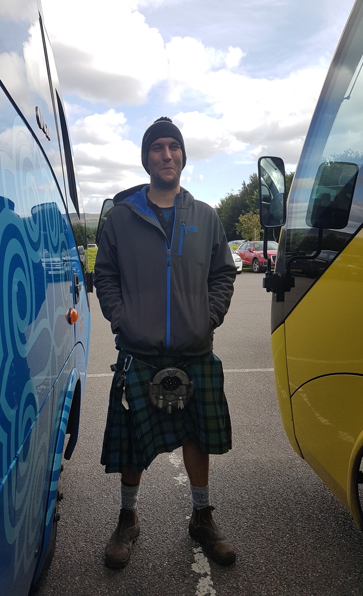 Un homme debout entre deux bus, portant un kilt et une veste.