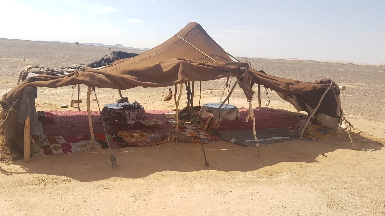 A traditional Berber-style fabric tent with rugs and low tables stands in a barren desert plain.