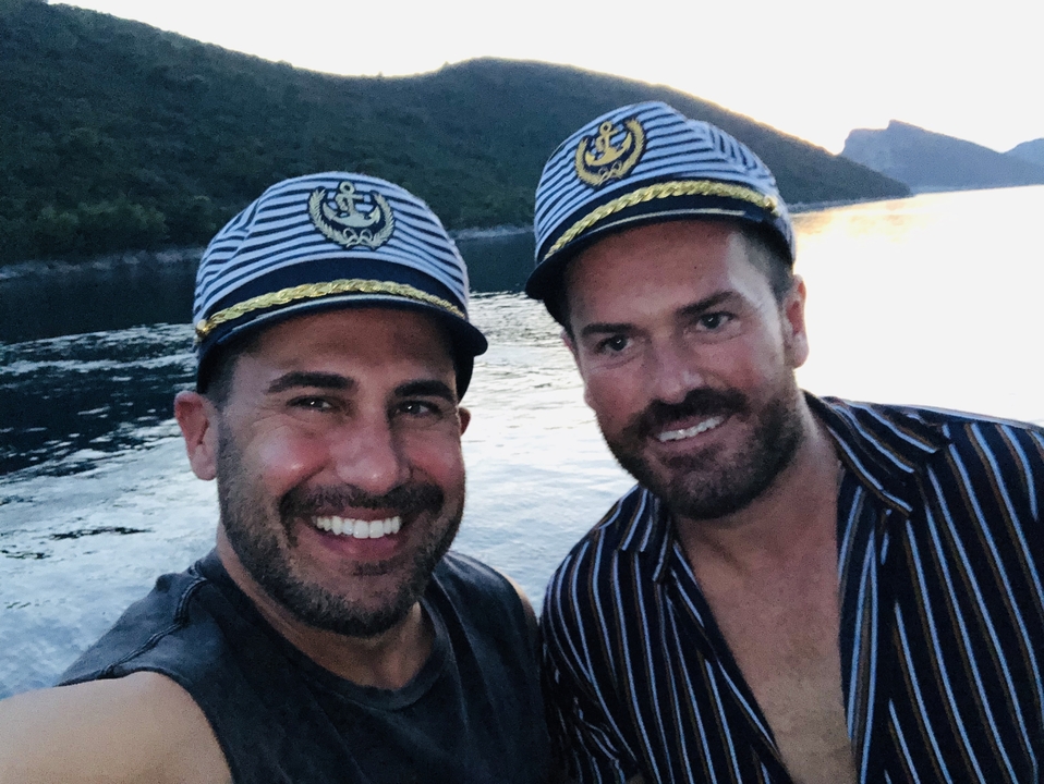 Deux hommes portant des chapeaux de marin qui sourient avec un paysage aquatique en arrière-plan.