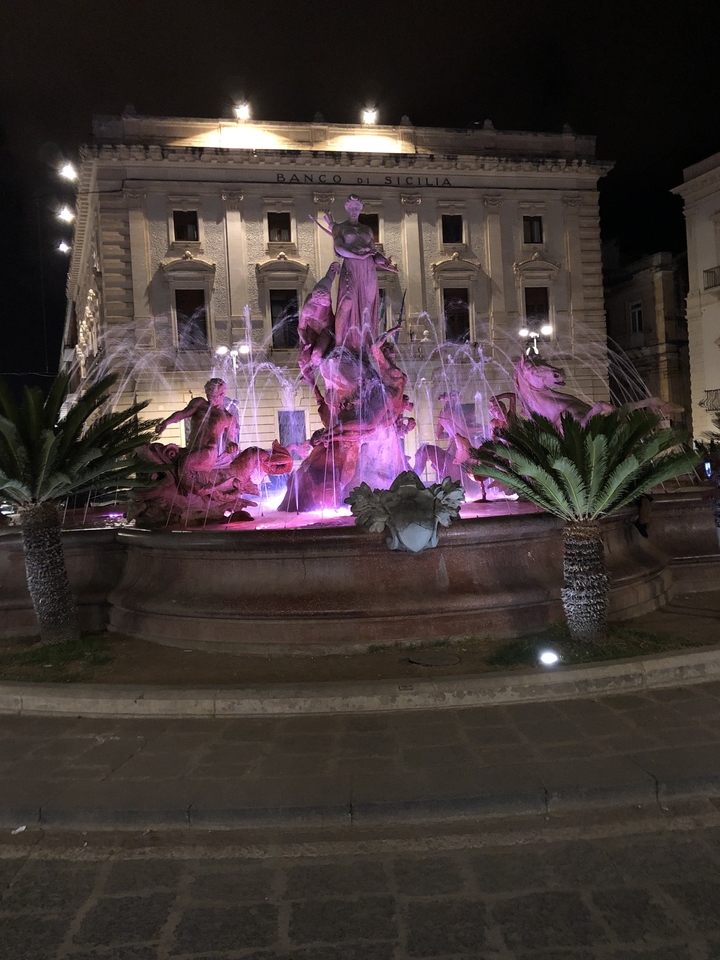 Fontaine illuminée avec sculptures et plantes autour.
