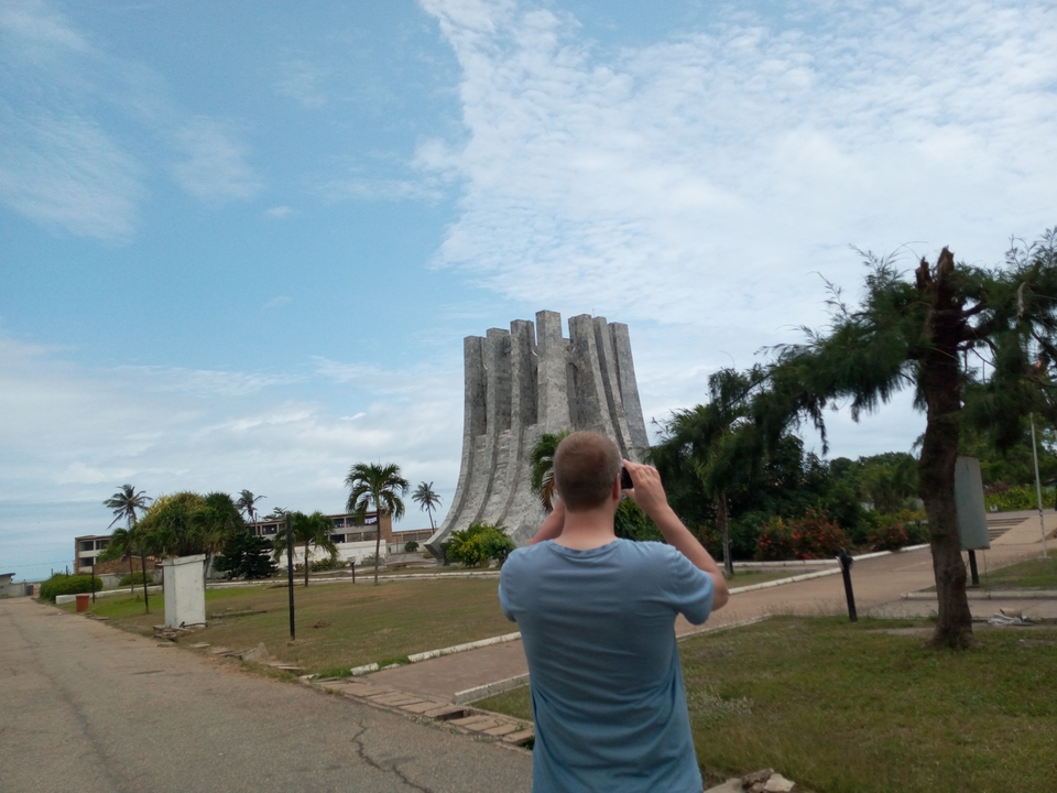 A visitor photographs a unique grey monument rising like stone petals amid palm trees in Ghana.