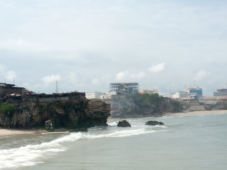 Hazy coastal view of rocky cliffs, crashing waves and urban buildings in the distance.