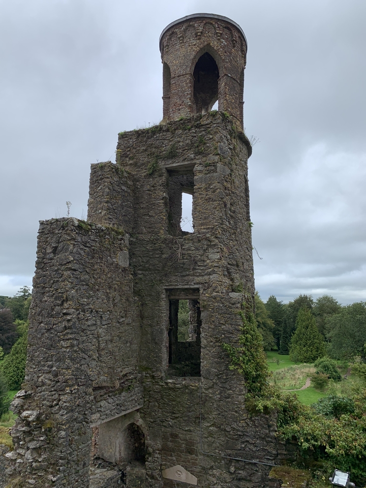 Une ruine de pierre avec des ouvertures rectangulaires entourée de verdure sous un ciel nuageux.