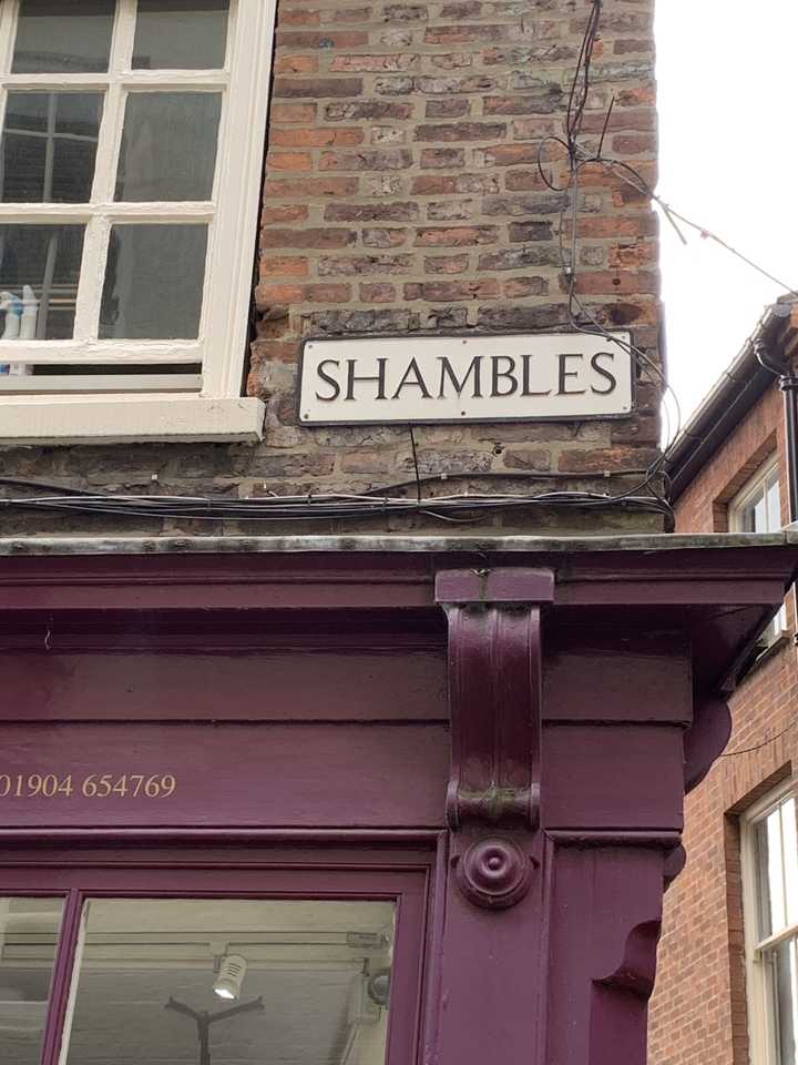 Panneau de rue sur un bâtiment en brique pour « Shambles ».
Note: "Shambles" is typically left untranslated when referring to the famous street name (like "The Shambles" in York, England), as it's a proper noun. If you need the word translated conceptually, "shambles" can mean "pagaille" or "désordre" in French, but for a street sign, the original name would usually be preserved.
