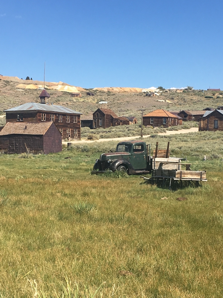 Vieux bâtiments rustiques et un véhicule abandonné dans un décor de ville fantôme.