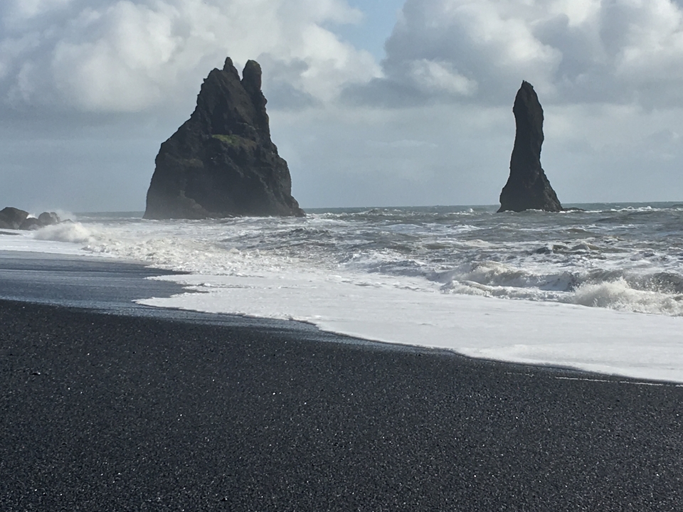 Plage de sable noir avec des aiguilles rocheuses et des vagues qui se brisent.
