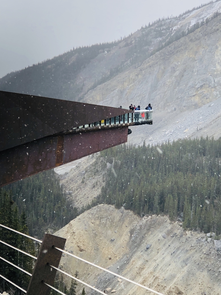 Eine Gruppe von Menschen, die auf einer Glasbrücke stehen und eine felsige Berglandschaft überblicken.