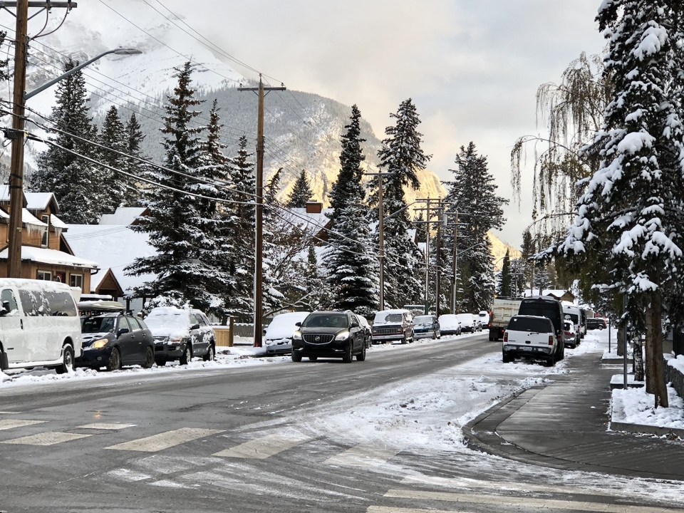 Eine verschneite Straße mit geparkten Autos und gehenden Menschen, gesäumt von Bäumen und Bergen im Hintergrund.