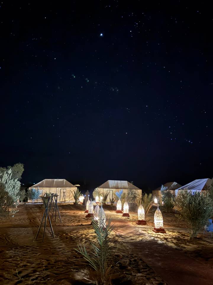 Scène nocturne d'un campement du désert sous un ciel étoilé avec des sentiers éclairés et des tentes.