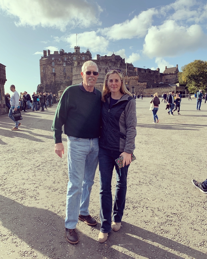 Deux personnes posant devant un bâtiment historique avec d'autres personnes en arrière-plan.