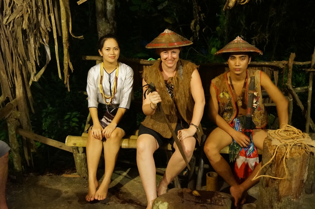 Deux personnes en tenue traditionnelle assises avec un touriste dans un cadre culturel.