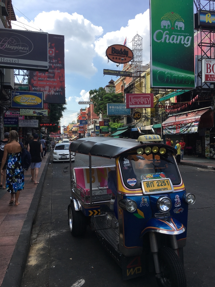 Scène de rue animée avec un tuk-tuk et diverses enseignes de magasins.