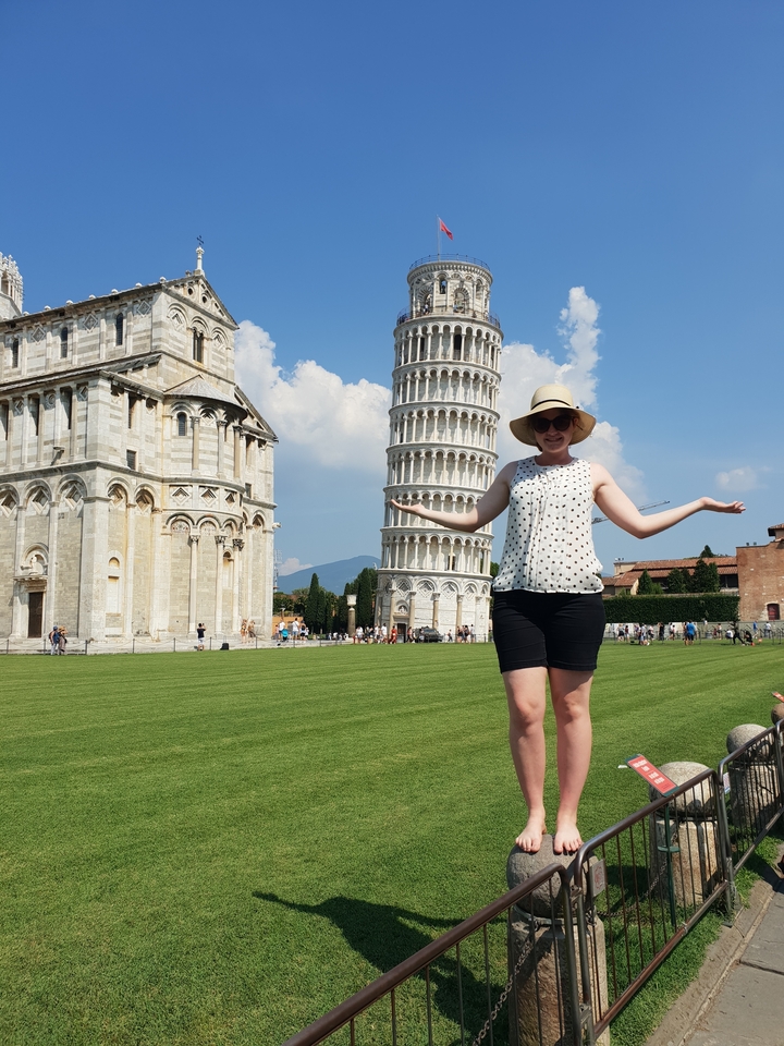 Une femme debout devant la Tour de Pise, les bras étendus.
