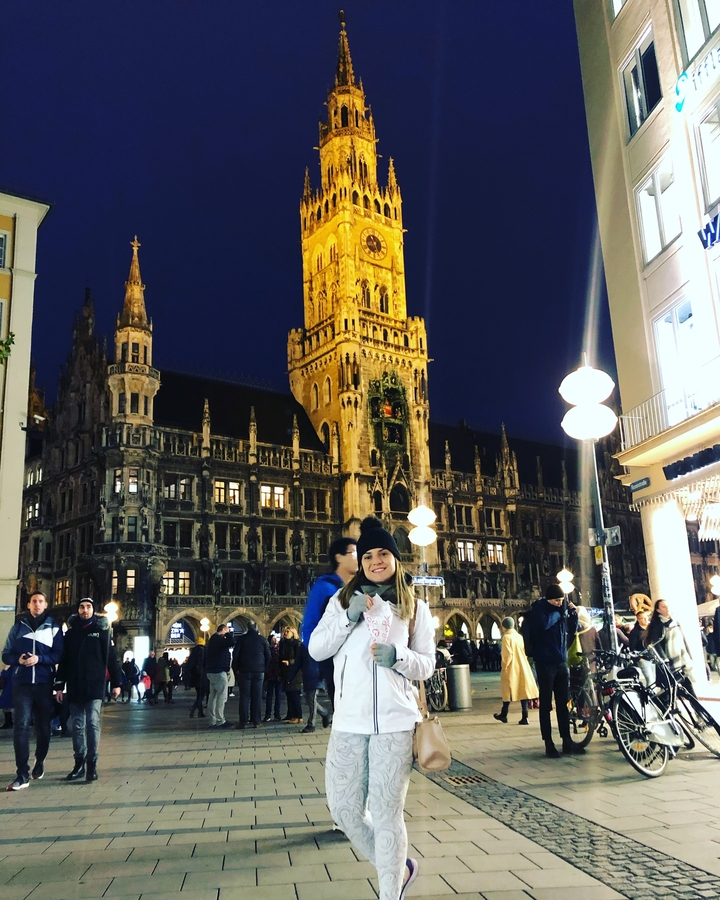 A person posing in front of the illuminated Rathaus-Glockenspiel at night.