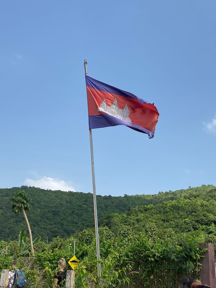 Drapeau cambodgien flottant contre un ciel bleu avec des collines vertes en arrière-plan.