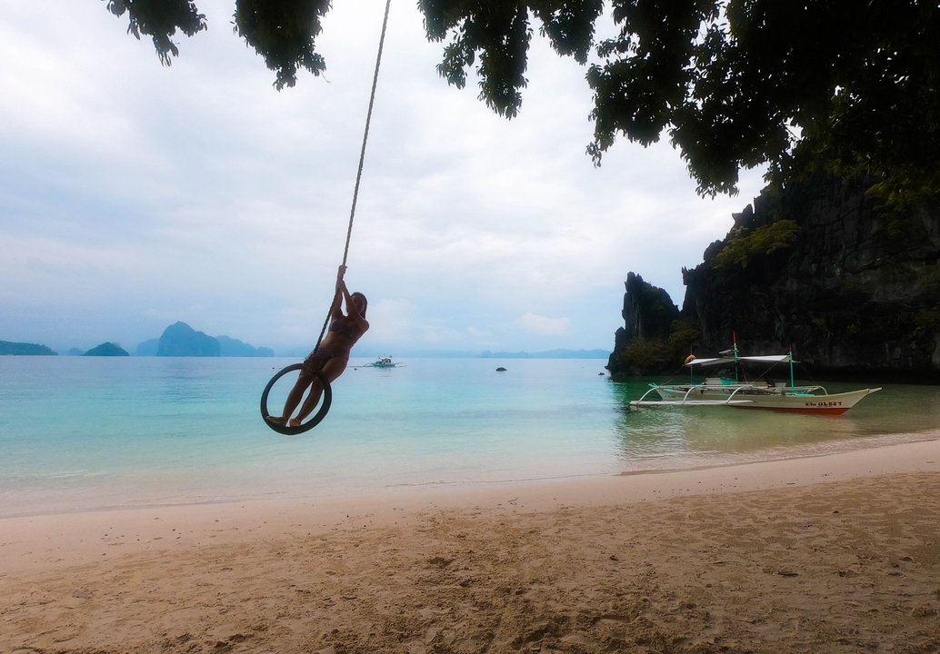 Personne se balançant sur une balançoire en corde à une plage avec des bateaux et des falaises en arrière-plan.