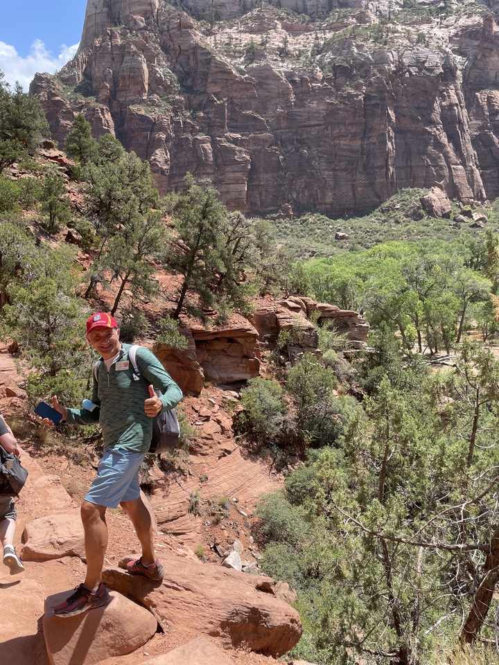 Randonneur montrant le pouce levé sur un sentier avec un paysage rocheux et de la végétation.