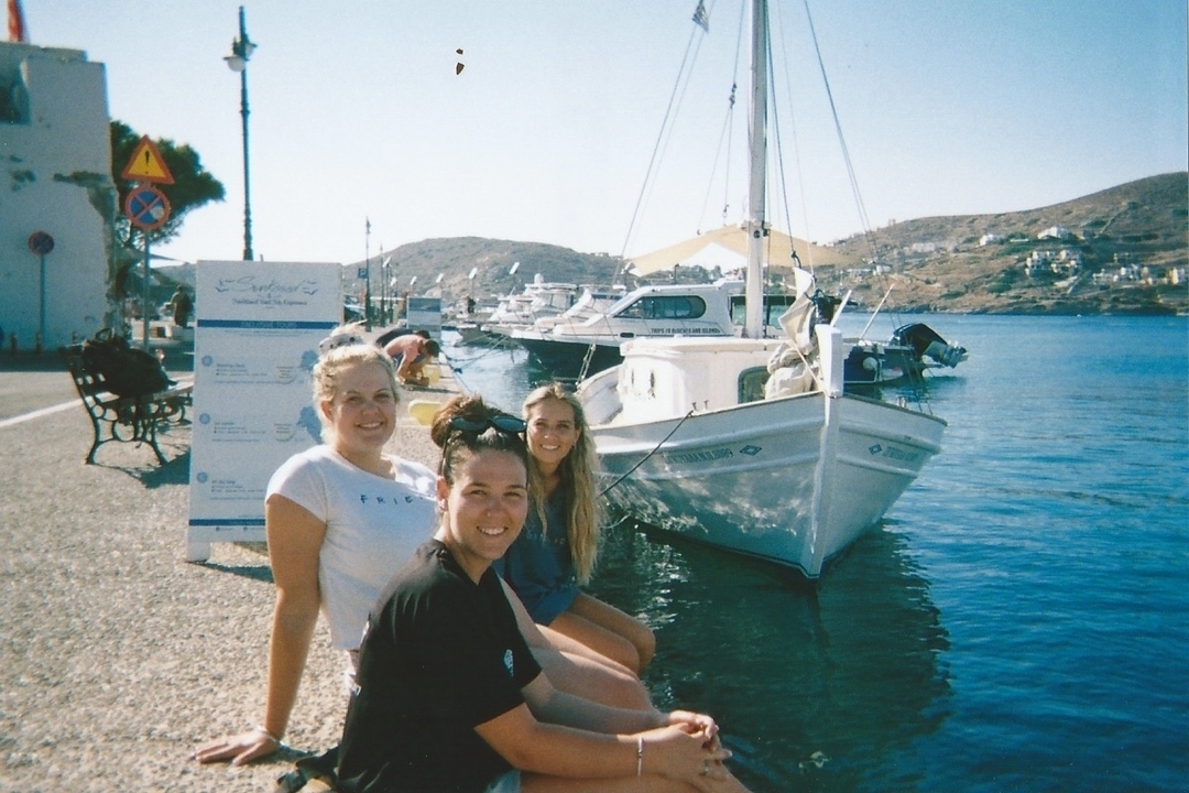 Three women sitting by the harbor with boats in the background.