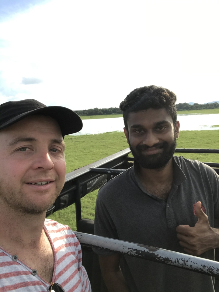 Deux personnes souriantes dans une zone herbeuse, lac en arrière-plan.