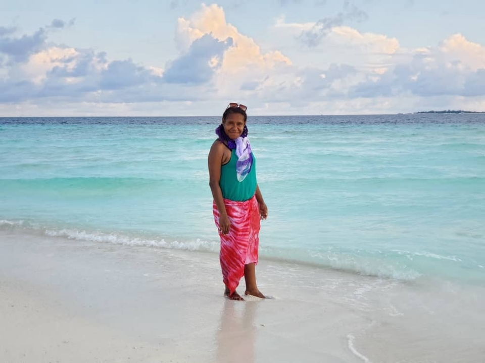 Femme debout sur une plage de sable avec de l'eau turquoise et un ciel nuageux en arrière-plan.