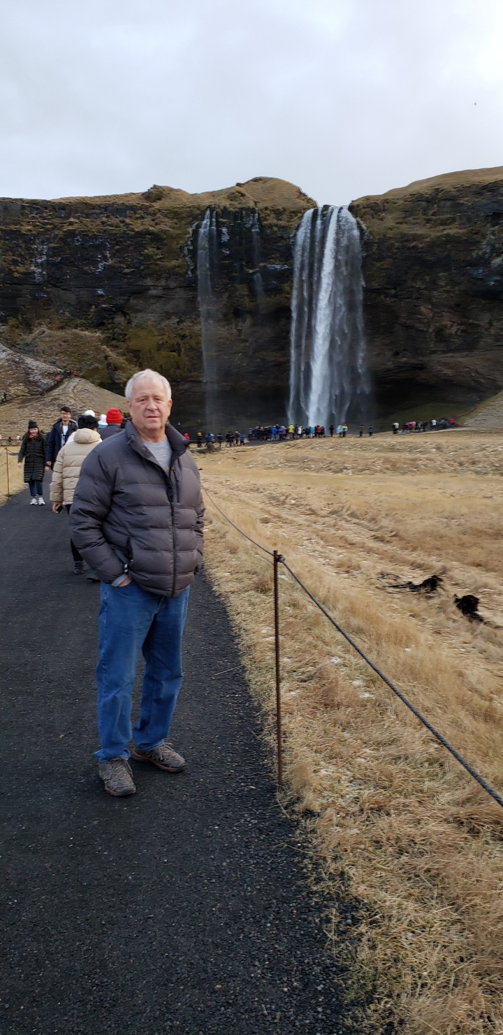 Man standing on a path with people in front of a majestic waterfall.