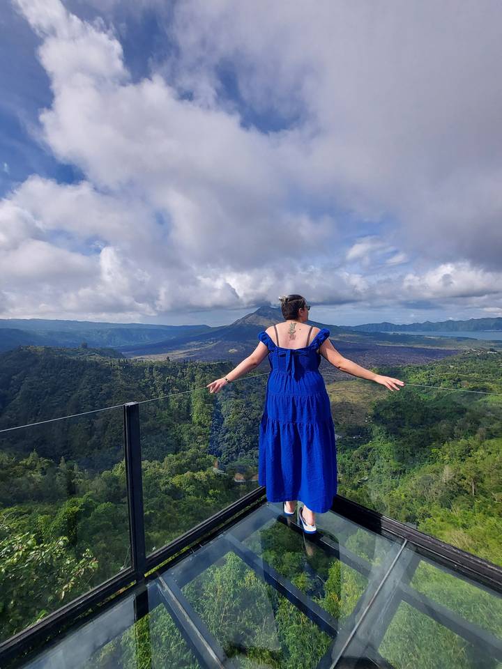 Une femme en robe bleue debout sur une plateforme de verre surplombant une forêt.