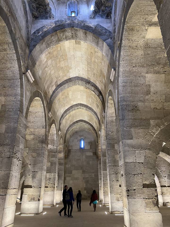 Intérieur d'une grande structure en pierre avec des plafonds voûtés et quelques personnes qui marchent.