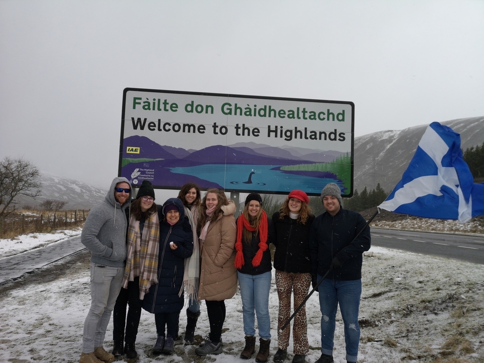 Groupe de personnes debout près d'un panneau souhaitant la bienvenue dans les Highlands avec des montagnes enneigées.