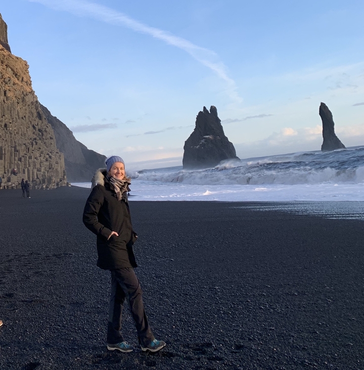 Femme sur une plage de sable noir avec des formations rocheuses dans l'océan.