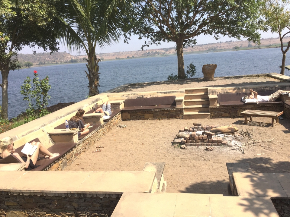 Travellers lounging on stone benches around sandy firepit terrace beside a large lake.