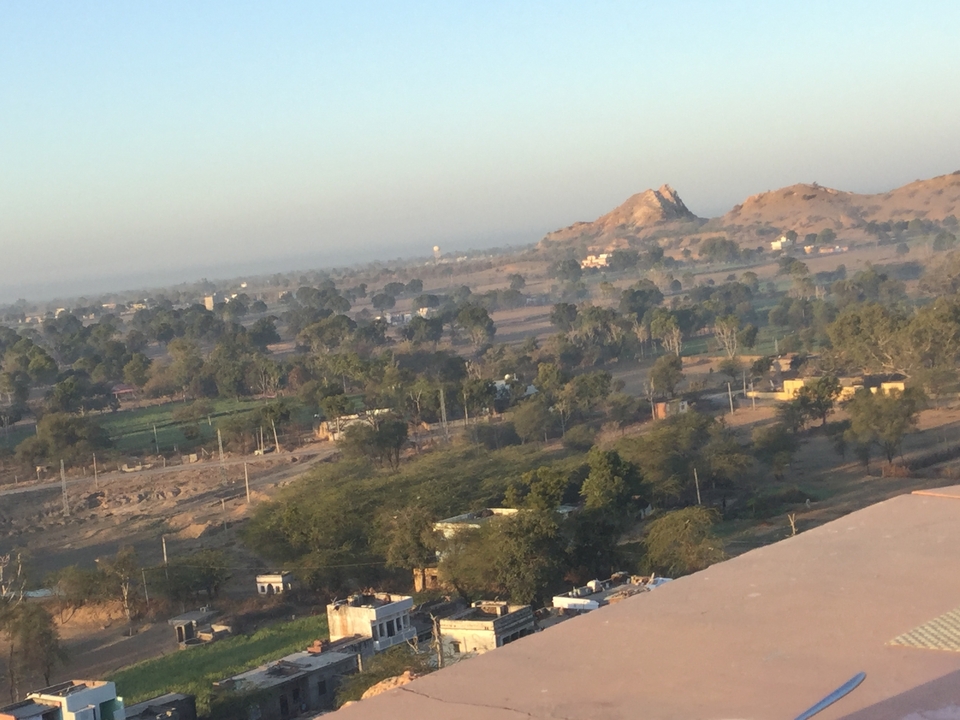 Hazy rural landscape of Rajasthan with scattered farms, scrub trees and distant rocky hill at dawn.