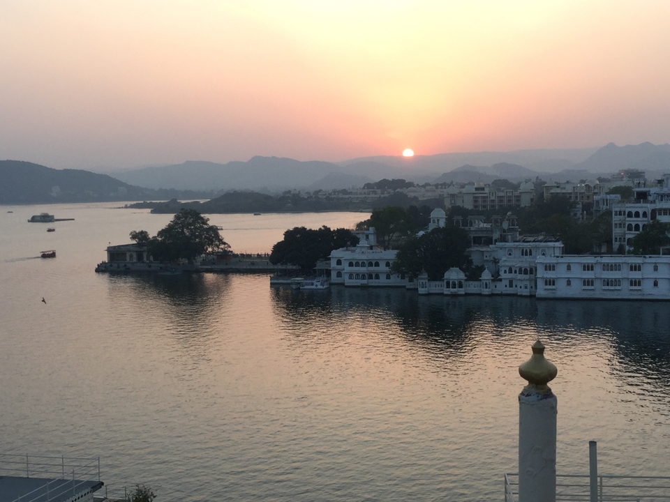 Sun setting over Lake Pichola with city palaces, boats and surrounding Aravalli hills in Udaipur.
