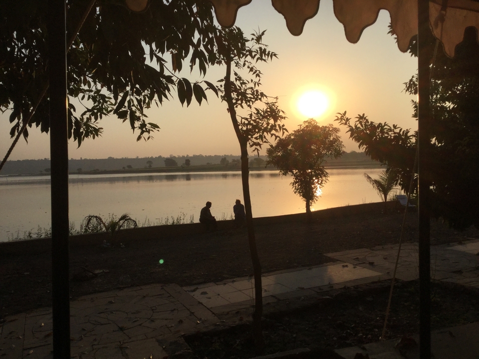 Low sun reflecting on tranquil lake while two figures sit on the shore beneath silhouetted trees.