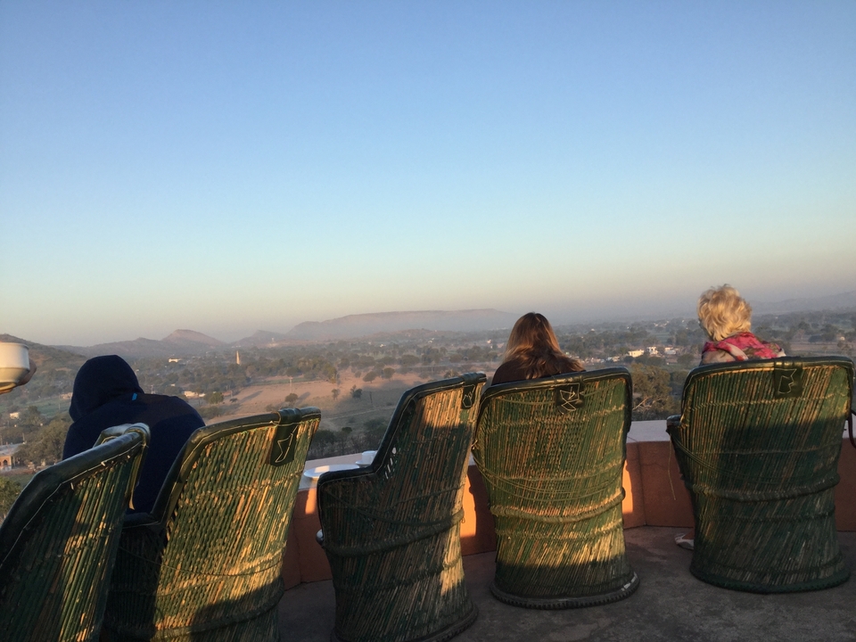 Travellers wrapped in blankets sit on wicker chairs atop fort ramparts overlooking misty countryside at dawn.