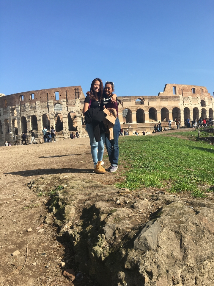Pair of travelers stand on grassy ground with the Colosseum stretching behind them