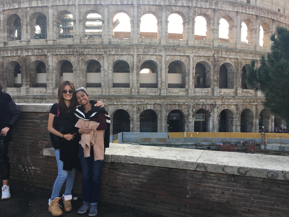 Two travelers pose arm-in-arm with the Roman Colosseum towering behind