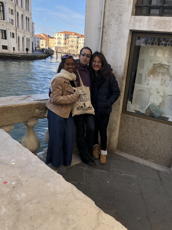 Trio of friends stand by a Venetian canal at dusk, water lapping against the stone balustrade