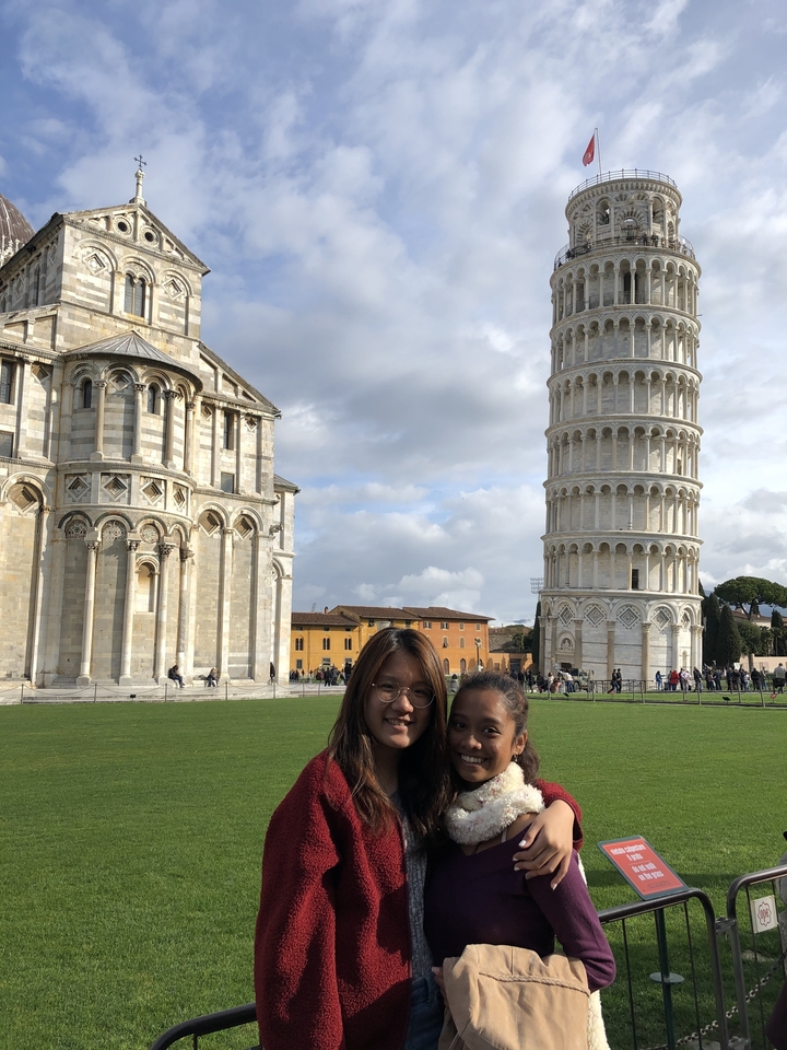 Two smiling travelers with the Leaning Tower of Pisa rising prominently behind them
