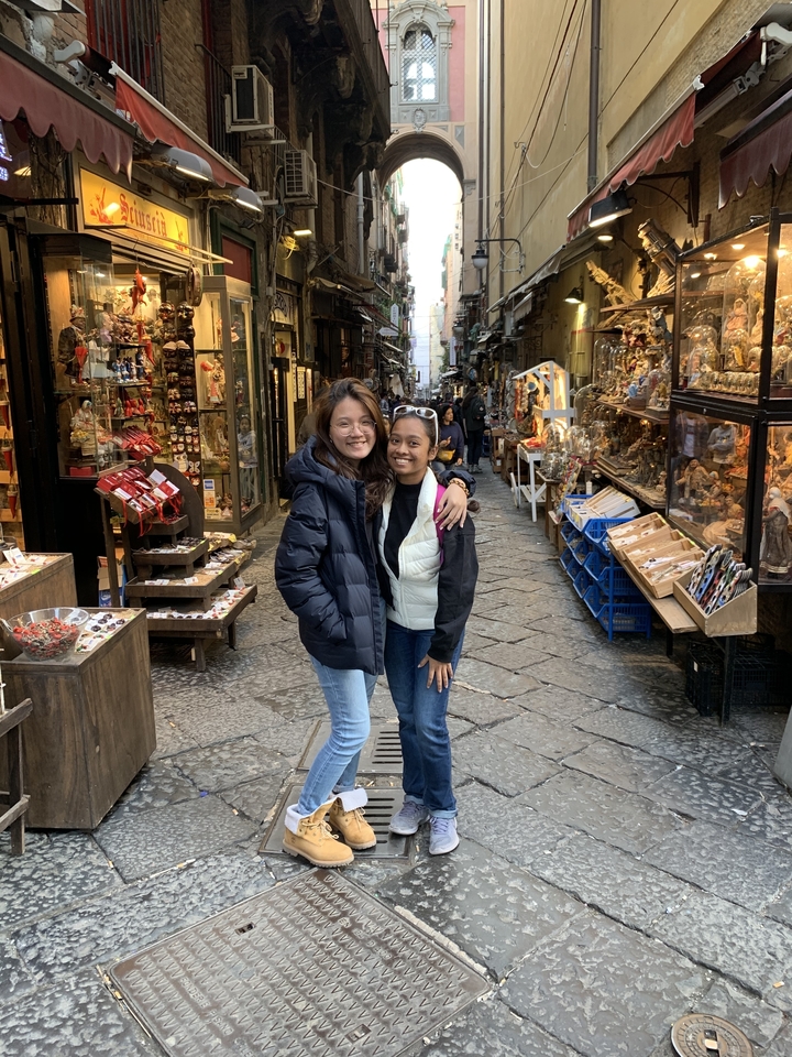 Two friends smile in a narrow cobblestone shopping lane filled with artisan stalls