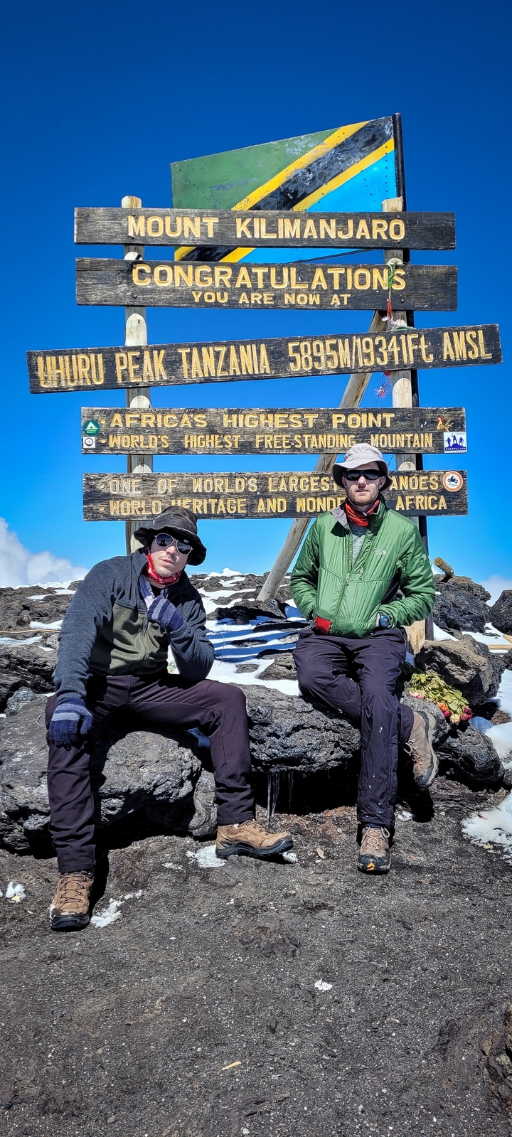 Deux personnes posant au sommet enneigé avec des panneaux indiquant la plus haute montagne isolée du monde.